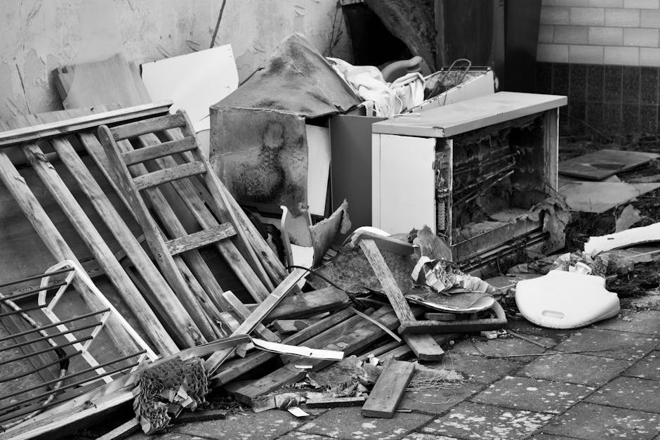 An outdoor scene depicting a pile of bulky waste and debris, including wooden slats, broken furniture, and miscellaneous household items, scattered on a paved surface. The area appears cluttered with various materials such as chipped wood, a desiccated appliance, and torn packaging, indicating waste awaiting disposal. A brick wall and part of a building are visible in the background, with natural lighting casting shadows over the mess. The surface shows signs of dirt and dust accumulated from the debris, and some items appear weathered and damaged, suggesting they have been abandoned outdoors. This image captures the necessity of thorough cleaning and waste removal services, which Cleaner Paddington offers as part of their expert cleaning and waste management solutions in Paddington W2.