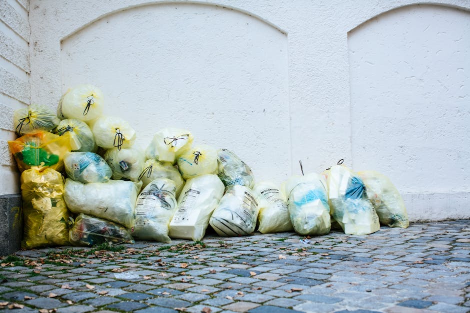 A collection of yellow, white, and blue garbage bags filled with bulky waste are piled up against a white exterior wall on a cobblestone pavement. The bags appear to contain various household refuse and are tightly sealed with black ties. The surface of the cobblestones shows some scattered leaves and small debris, indicating outdoor conditions. The background features a plain white wall with a slight texture, reflecting natural daylight. The scene illustrates waste disposal steps typical of commercial or domestic cleaning services in Paddington, with Cleaner Paddington providing professional bulky waste clearance and disposal in W2 area.