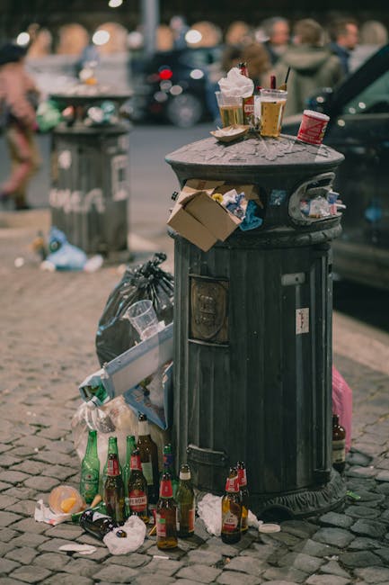 An outdoor scene depicting a pile of bulky waste and debris, including wooden slats, broken furniture, and miscellaneous household items, scattered on a paved surface. The area appears cluttered with various materials such as chipped wood, a desiccated appliance, and torn packaging, indicating waste awaiting disposal. A brick wall and part of a building are visible in the background, with natural lighting casting shadows over the mess. The surface shows signs of dirt and dust accumulated from the debris, and some items appear weathered and damaged, suggesting they have been abandoned outdoors. This image captures the necessity of thorough cleaning and waste removal services, which Cleaner Paddington offers as part of their expert cleaning and waste management solutions in Paddington W2.
