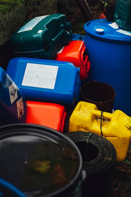 An outdoor scene featuring various hazardous waste containers in multiple colors such as blue, yellow, red, and black, placed on a surface with grass and foliage in the background. The containers are made of durable plastic, with some showing labels or warnings related to hazardous materials. A few containers have visible caps or lids, and one black container appears open. The setting suggests an area designated for waste storage before proper disposal, with natural lighting illuminating the scene. This arrangement reflects proper handling and storage practices for hazardous waste, which is relevant to the context of legal steps for hazardous waste removal in Paddington, as emphasized by Cleaner Paddington, a professional cleaning service specializing in environmental hygiene and waste management.