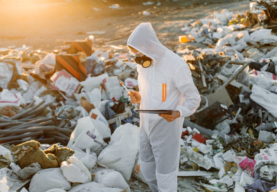 A person dressed in white protective coveralls, wearing a hood, gloves, and a respirator mask, standing amidst a large pile of mixed waste and debris on a sandy beach during sunset. The individual is holding a clipboard in one hand and a smartphone in the other, seemingly assessing or documenting the hazardous waste. The scene highlights the importance of proper hazardous waste management and cleanup, as part of responsible environmental hygiene and sanitisation efforts, relevant to the context of legal steps for hazardous waste removal in Paddington. The background features a sunset sky, casting a warm glow over the scattered waste and the worker engaged in environmental safety protocols.