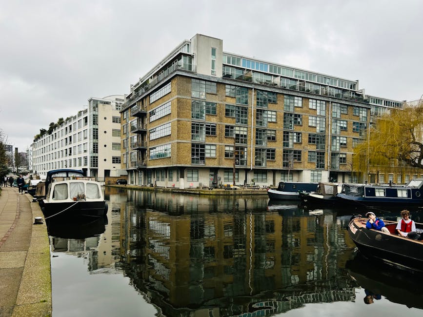 A photograph of a peaceful canalside scene in Little Venice, Paddington, taken during daytime with bright natural lighting. The calm water reflects the blue sky with scattered clouds and is bordered b
