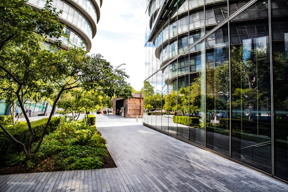 The image shows a modern office building with a curved glass facade reflecting the surrounding greenery and sky, located at Paddington Basin. In front of the building, there is a wide, clean paving walkway with smooth, light gray stone tiles. To the left, well-maintained landscaping includes a small tree with lush green leaves and surrounding bushes. The sunlight highlights the shiny surface of the glass, emphasizing its cleanliness and transparency. Pedestrians are visible walking in the background, and the overall scene conveys a tidy, professional environment consistent with commercial premises. The reflection in the glass showcases further garden spaces and a bright, partly cloudy sky, suggesting good lighting conditions. Cleaner Paddington, a reputable cleaning company, emphasizes the importance of maintaining the pristine appearance of commercial office spaces through thorough cleaning and sanitisation routines.