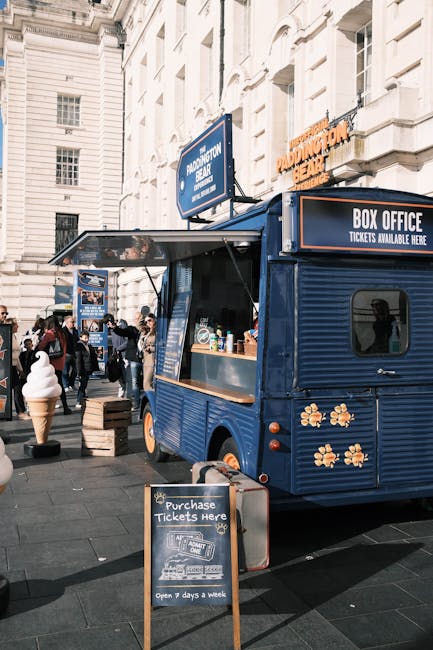 A blue mobile food stand with a corrugated metal exterior featuring decorative flower decals is positioned on a busy street in Paddington. The stand has a large service window displaying various supplies and a small chalkboard sign in front advertising ticket purchases, indicating it operates seven days a week. Surrounding the stand are pedestrians, some in motion and others engaged in conversation. Nearby, a large ice cream model on a stand and additional street furniture are visible, with classic white stone building facades and windows in the background. The scene is captured during daylight hours, with natural lighting highlighting the cleanliness and arrangement of the area, complemented by a visible signage indicating the location as Paddington Basin. Cleaner Paddington is committed to maintaining hygienic and well-presented outdoor environments, ensuring surfaces such as the stand’s exterior and surrounding pavement remain clean and free of dust or dirt, reflecting their expertise in surface cleaning and sanitation for commercial spaces.