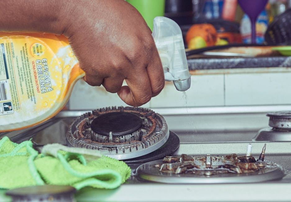 A close-up view of a person's hand holding a spray bottle, applying cleaning solution onto a gas stove burner in a kitchen. The stove appears to be used frequently, showing visible grease and grime around the burner. A green cleaning cloth is draped nearby on the kitchen countertop, which is made of a smooth, light-colored material. The background includes some colorful objects and utensils, indicating a typical domestic kitchen setting. The overall scene emphasizes surface cleaning and deep cleaning processes essential for maintaining hygiene in a kitchen. The image reflects the professional cleaning services provided by Cleaner Paddington, highlighting their expertise in domestic cleaning and grease removal, especially relevant to emergency deep-clean tips for kitchen grease, as explored on the Paddington kitchen grease page.
