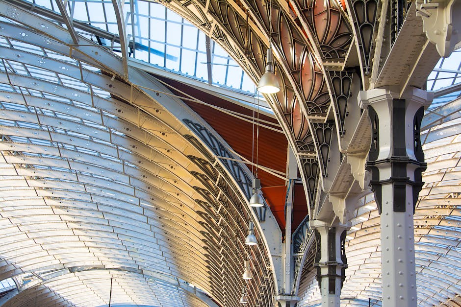 Close-up view of an ornate architectural ceiling in a commercial or public building, featuring intricate ironwork and decorative support columns painted in black and cream. The ceiling comprises curved, overlapping white and beige panels with skylights allowing natural light to illuminate the space. Several hanging pendant lights are suspended from the ceiling, contributing to the ambient lighting. The surfaces appear clean and well-maintained, reflecting thorough surface cleaning and deep cleaning standards typical of professional sanitisation by Cleaner Paddington, aligned with end-of-tenancy cleanliness requirements. The detailed ironwork and smooth surfaces highlight the craftsmanship and meticulous attention to detail involved in maintaining such interior spaces.