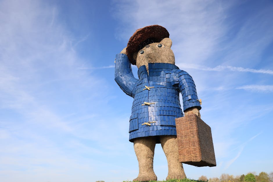 A large outdoor sculpture of a figure resembling Paddington Bear, characterized by its beige textured surface, wearing a blue coat with wooden toggle fastenings, a brown hat, and holding a brown suitcase in its right hand. The figure is gazing into the distance against a bright blue sky with wispy white clouds, situated on a grassy field. The sculpture's surfaces, especially the coat and hat, appear detailed with a textured effect, contrasting with the smoother background of the sky. The sunlight illuminates the statue, emphasizing its size and the craftsmanship in its construction, with shadows cast on the ground. This sculpture presents a whimsical, iconic representation of Paddington Bear in an outdoor setting.