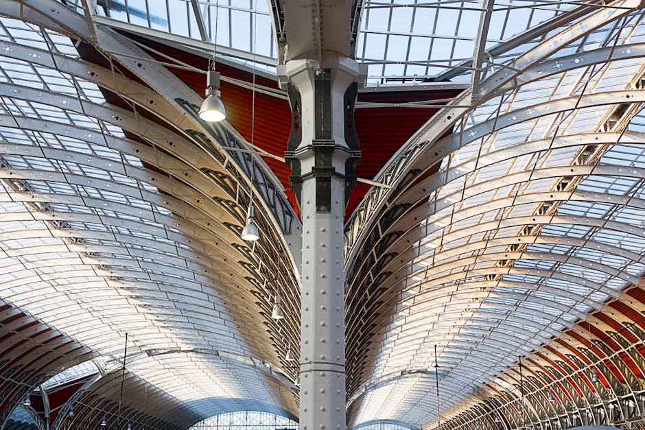 Close-up view of an ornate architectural ceiling in a commercial or public building, featuring intricate ironwork and decorative support columns painted in black and cream. The ceiling comprises curved, overlapping white and beige panels with skylights allowing natural light to illuminate the space. Several hanging pendant lights are suspended from the ceiling, contributing to the ambient lighting. The surfaces appear clean and well-maintained, reflecting thorough surface cleaning and deep cleaning standards typical of professional sanitisation by Cleaner Paddington, aligned with end-of-tenancy cleanliness requirements. The detailed ironwork and smooth surfaces highlight the craftsmanship and meticulous attention to detail involved in maintaining such interior spaces.
