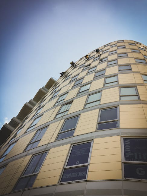 An upward-looking view of a curved, multi-story residential building with beige and light brown exterior cladding, featuring numerous rectangular windows, some of which display reflective glass. The building's smooth surface appears clean and well-maintained under natural daylight, with a clear blue sky in the background. The photograph emphasizes the modern architectural design, with no visible signs of dirt or grime. Cleaner Paddington specializes in surface cleaning and sanitisation services for residential properties, ensuring their exteriors remain spotless and preserved.
