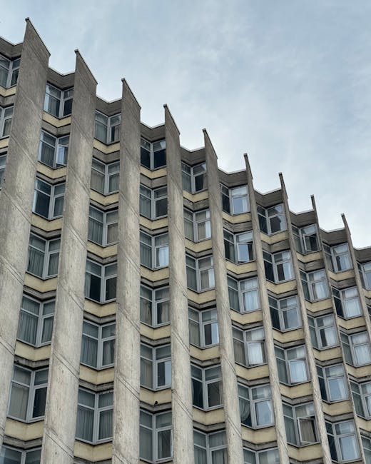 An upward-looking view of a curved, multi-story residential building with beige and light brown exterior cladding, featuring numerous rectangular windows, some of which display reflective glass. The building's smooth surface appears clean and well-maintained under natural daylight, with a clear blue sky in the background. The photograph emphasizes the modern architectural design, with no visible signs of dirt or grime. Cleaner Paddington specializes in surface cleaning and sanitisation services for residential properties, ensuring their exteriors remain spotless and preserved.