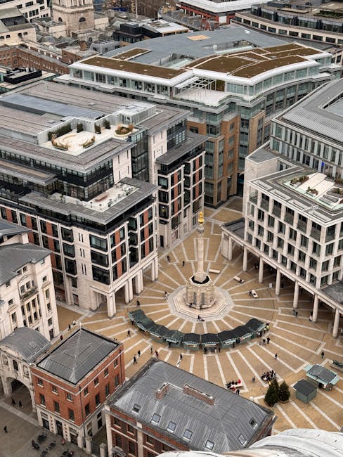 An aerial view of a modern urban plaza surrounded by multi-story commercial and residential buildings with a mix of glass and concrete facades. The plaza features a patterned stone surface with radial lines originating from a central monument, which is a tall stone obelisk on a square base. Benches and small planters are arranged around the monument, with a few pedestrians walking through the open space. The surrounding buildings have large windows, rooftop terraces, and some rooftop gardens, all illuminated by natural daylight. The scene captures the clean, structured look of a well-maintained city center, typical of high-quality cleaning and maintenance standards, as promoted by Cleaner Paddington for commercial and residential surfaces.