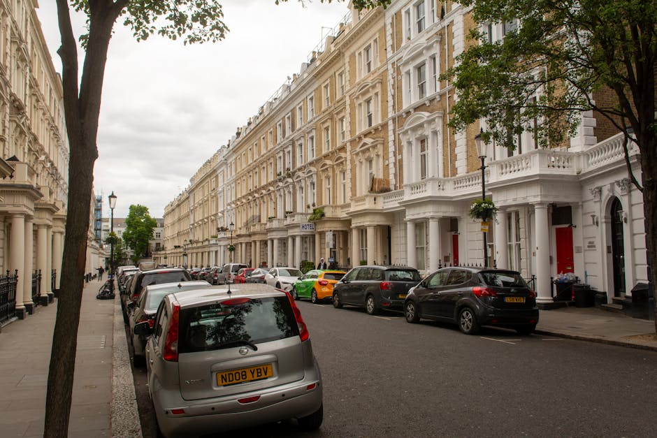 A residential street in Paddington featuring several white Victorian-style terraced buildings with ornate architectural details, large sash windows, and decorative facades. The street is lined with parked cars of various colors and models along the curb, with some trees providing greenery and shade. The pavement is clean and well-maintained, with lampposts and a hotel sign visible in the background, suggesting a tidy and well-kept urban environment. The lighting indicates an overcast day, highlighting the street's orderly appearance. This image reflects the typical residential area where professional cleaning services by Cleaner Paddington may be employed to maintain hygiene and appearance.