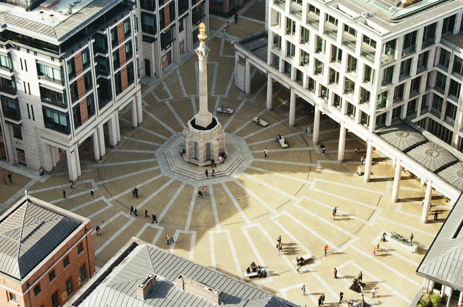 An aerial view of a modern urban plaza surrounded by multi-story commercial and residential buildings with a mix of glass and concrete facades. The plaza features a patterned stone surface with radial lines originating from a central monument, which is a tall stone obelisk on a square base. Benches and small planters are arranged around the monument, with a few pedestrians walking through the open space. The surrounding buildings have large windows, rooftop terraces, and some rooftop gardens, all illuminated by natural daylight. The scene captures the clean, structured look of a well-maintained city center, typical of high-quality cleaning and maintenance standards, as promoted by Cleaner Paddington for commercial and residential surfaces.